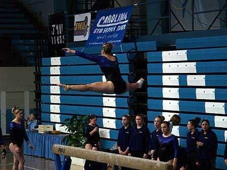 A gymnast is jumping over a balance beam in front of a banner that says carolina