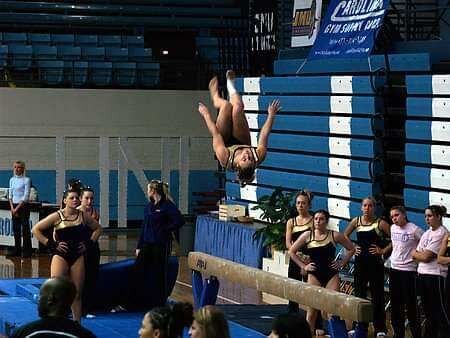 A female gymnast is doing a trick on a balance beam while a group of people watch.