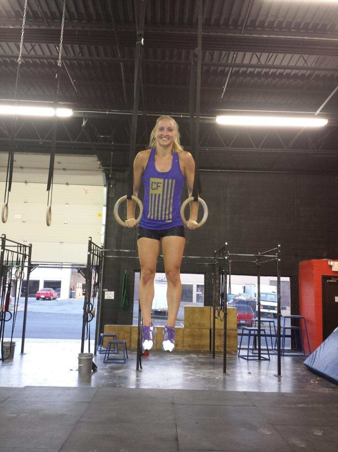 A woman in a purple tank top is hanging from gymnastic rings