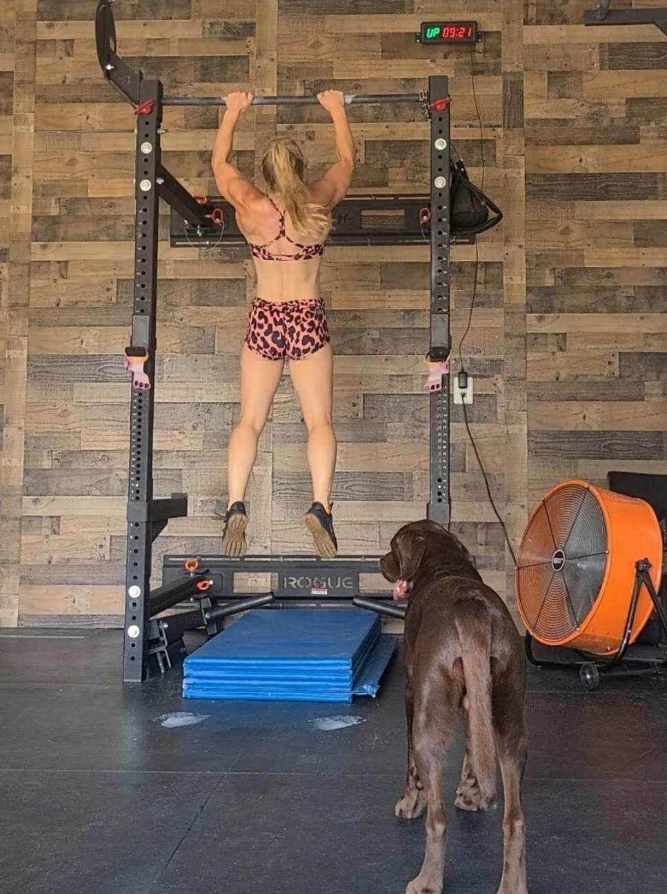 A woman is doing pull ups in a gym while a dog watches.