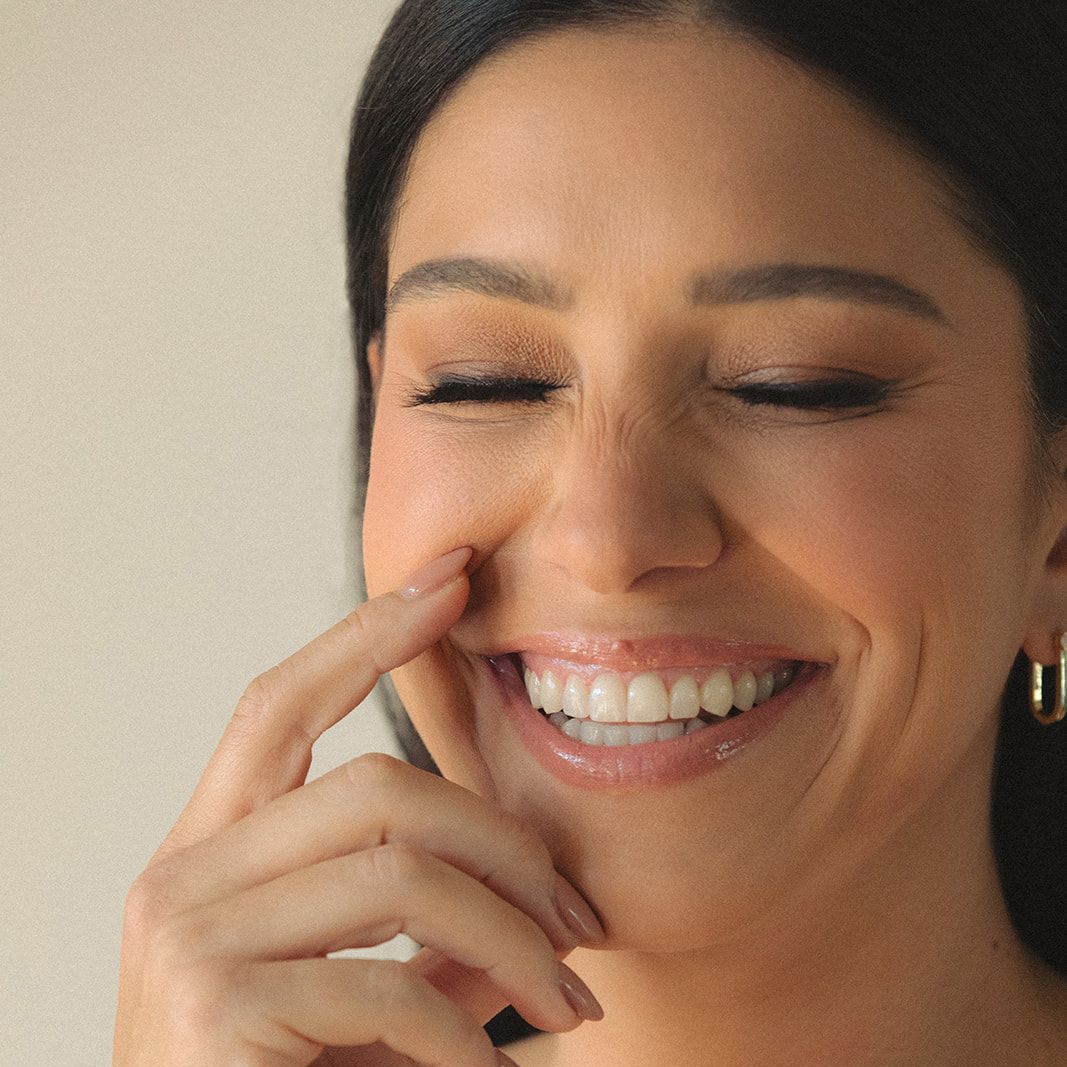 Woman smiling, touching her cheek with a finger. She has fair skin, dark hair, and is wearing makeup.