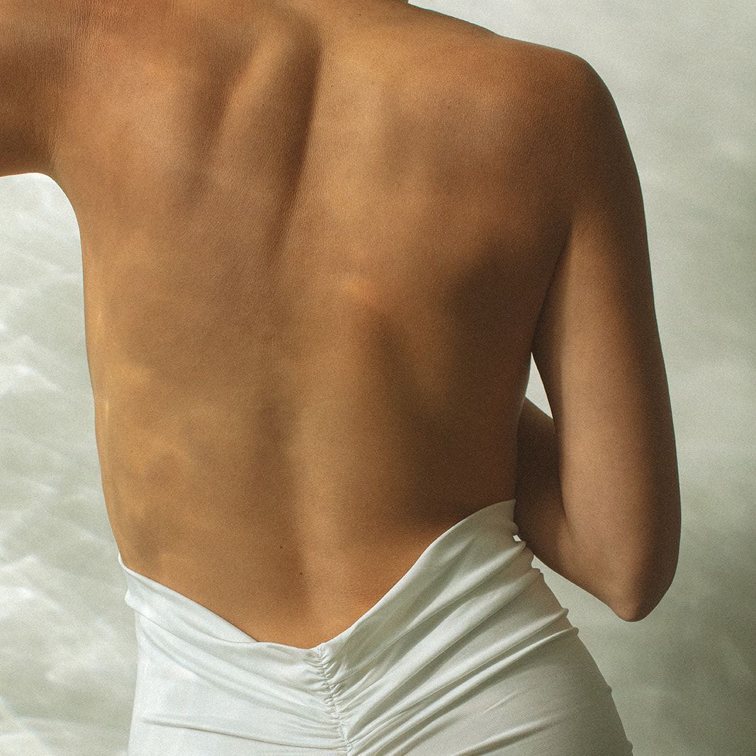A person's back, in a white dress, with their arms bent; showing a tan skin tone.