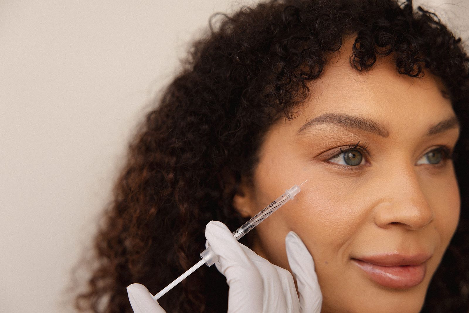 A gloved hand uses a syringe to administer an injection under the eye of a person, who looks forward with a neutral smile.