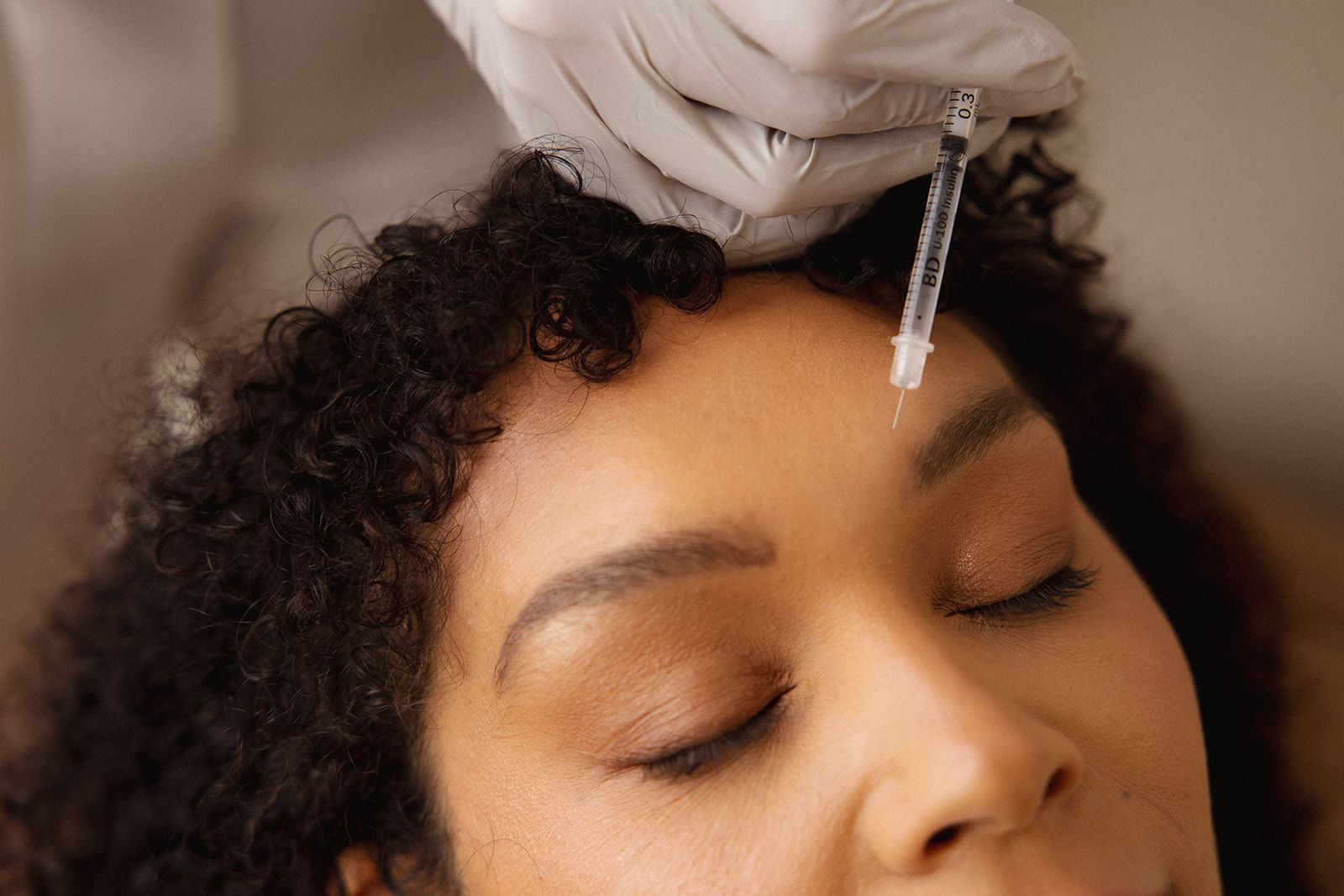 A gloved hand holds a needle near a person's forehead, preparing for a cosmetic injection.