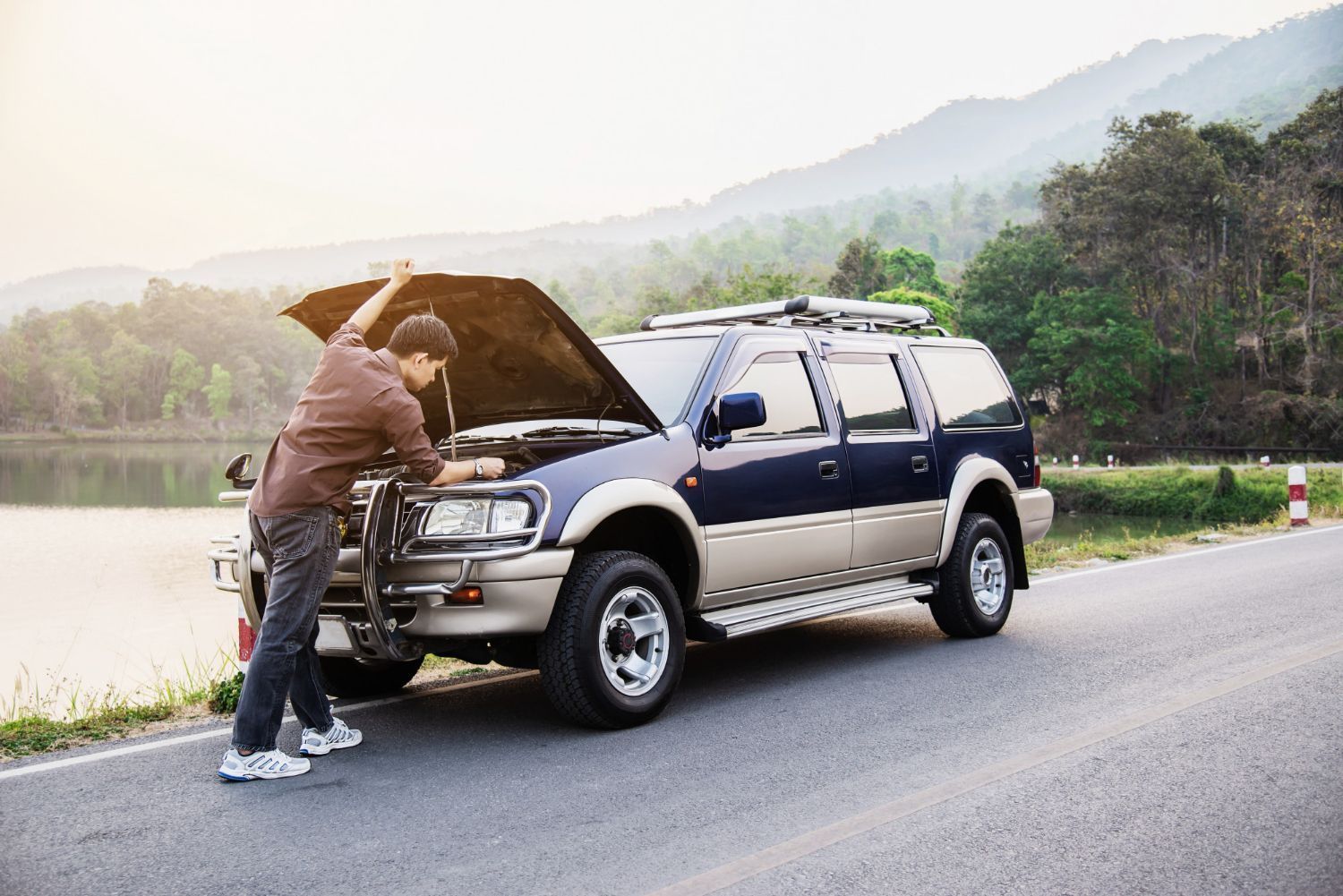 Man standing by broken-down blue SUV with hood open on side of road near lake and mountains.