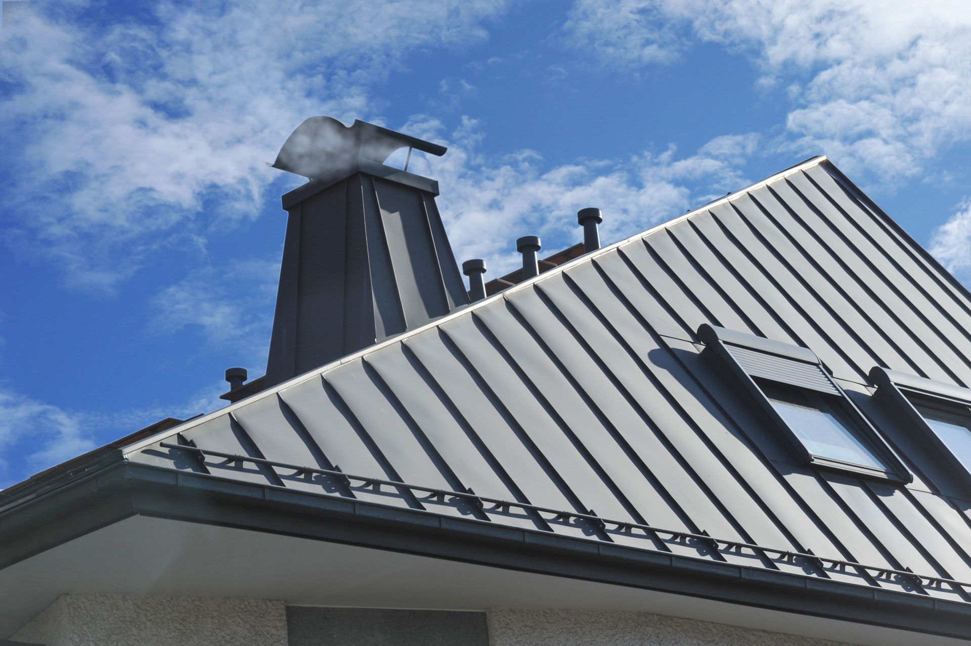 Gray metal roof with chimney and skylight against a cloudy blue sky.