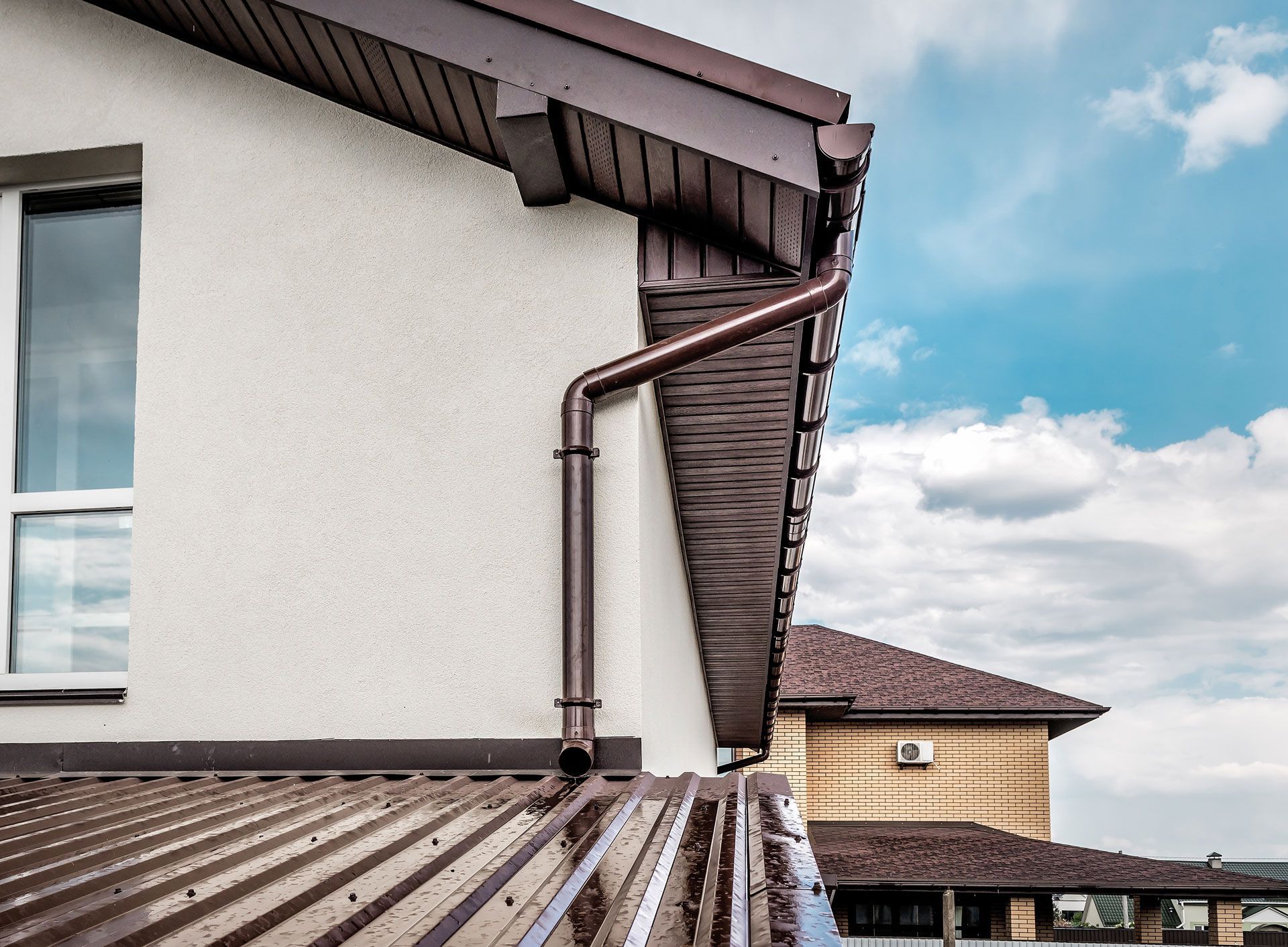Brown gutters on a beige house with a window. Another house with a brown roof in the background.