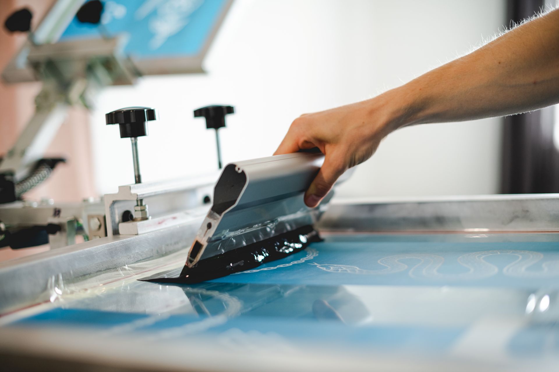 Hands using a squeegee to apply red ink through a screen print frame onto a black shirt with a white skull design.