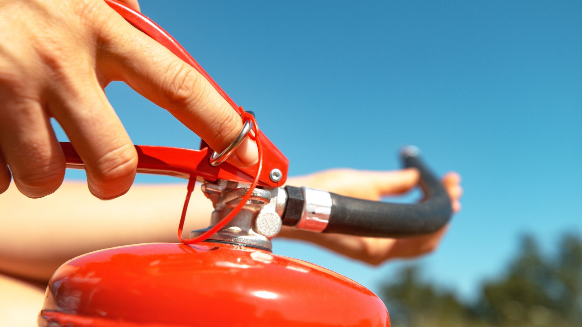 A person presses the handle of a red fire extinguisher while aiming the hose outdoors.