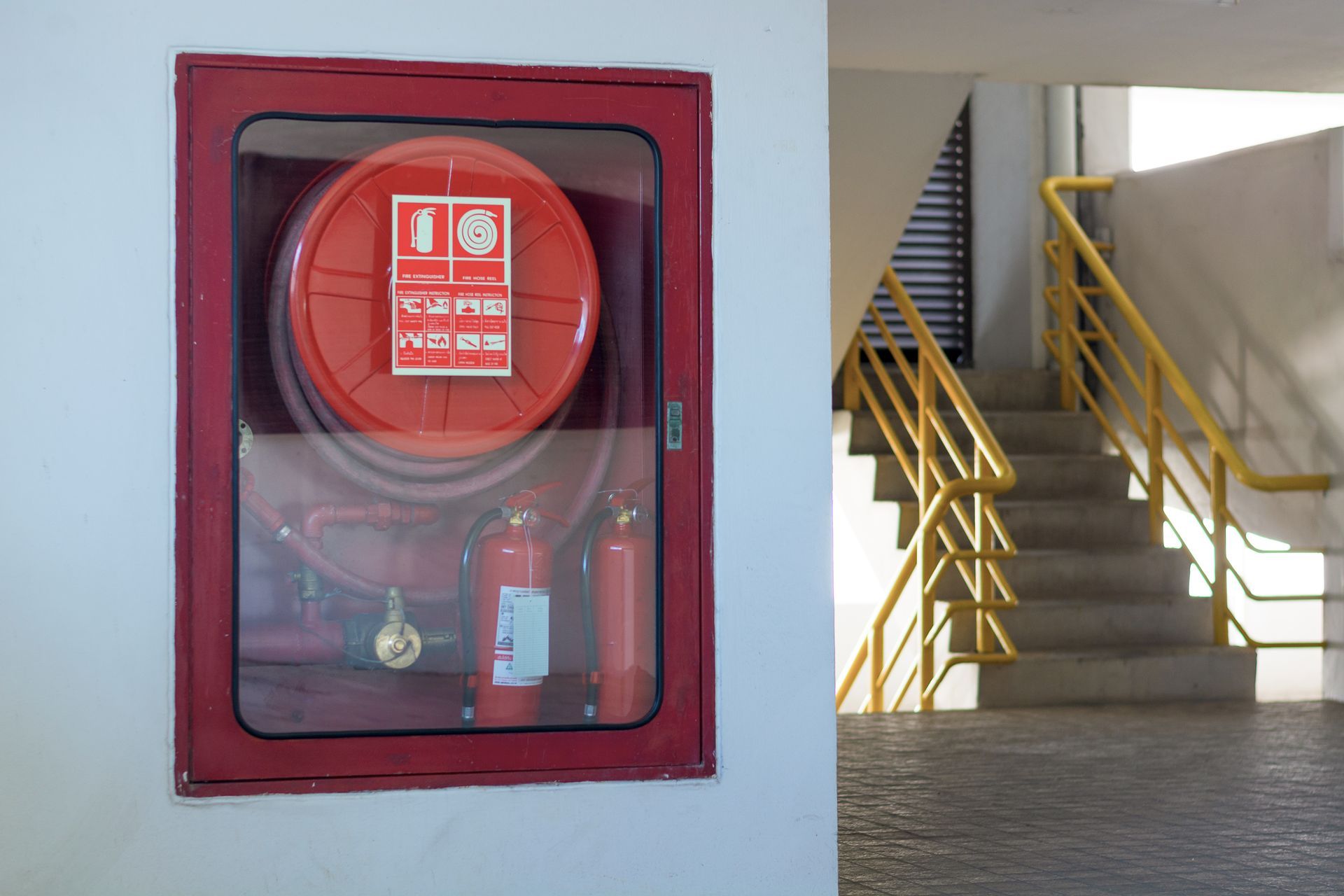 Red fire hose cabinet and extinguishers by yellow stair railings on wall.