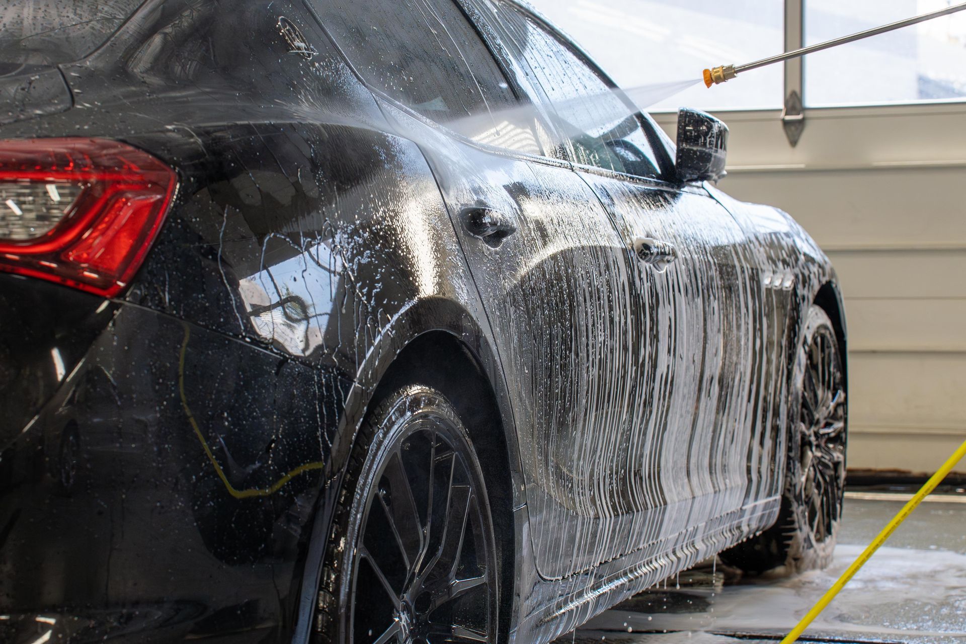 Black car being washed at an indoor car wash, covered in soap suds.