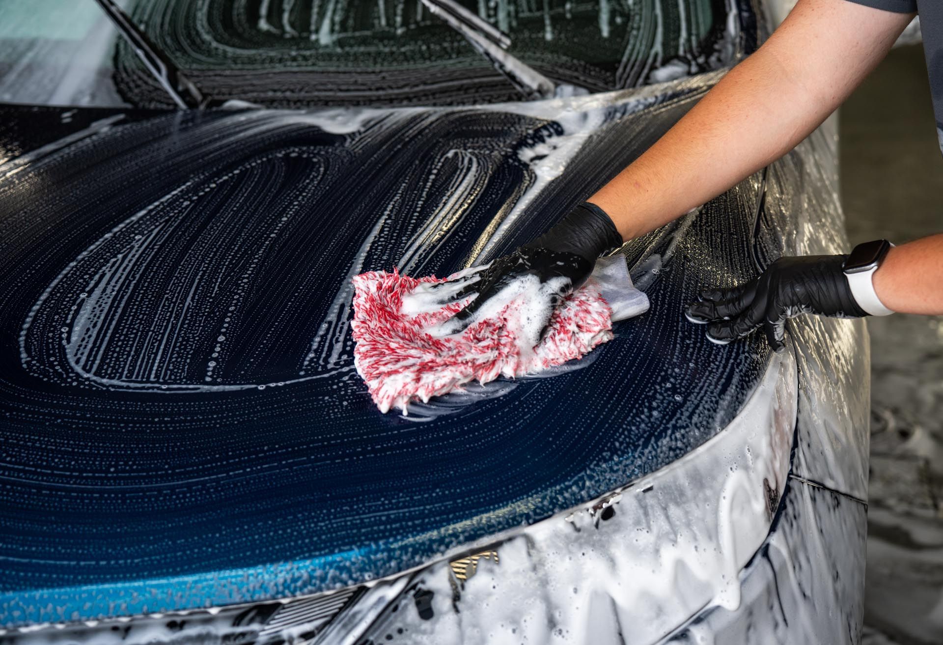 Gloved hands scrubbing a foam-covered black car hood with a red sponge