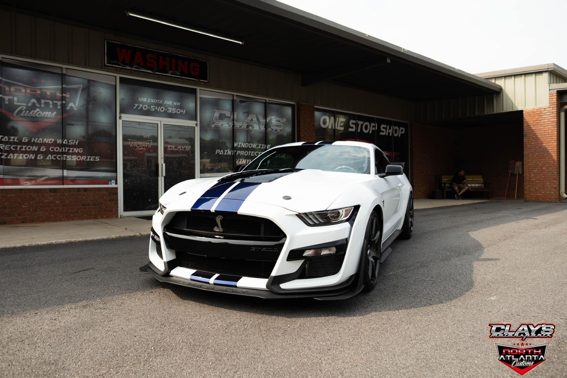 A white mustang with blue stripes is parked in front of a building.