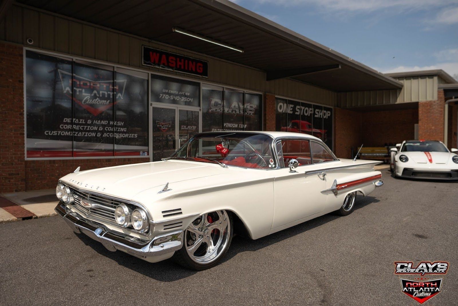 Classic white lowrider parked outside a storefront, with a white car in the background.