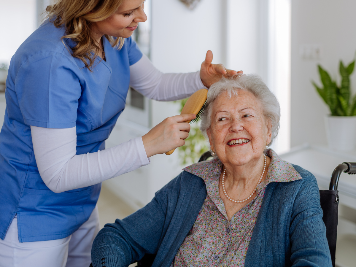 Caregiver brushing the hair of an elderly woman in a wheelchair, both smiling. Indoors.