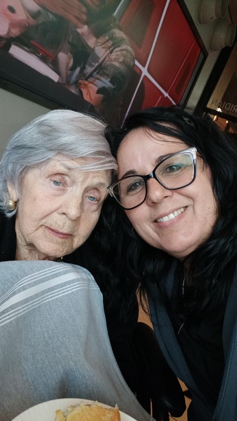 Woman with glasses smiles next to an older woman. Both are close, indoors.