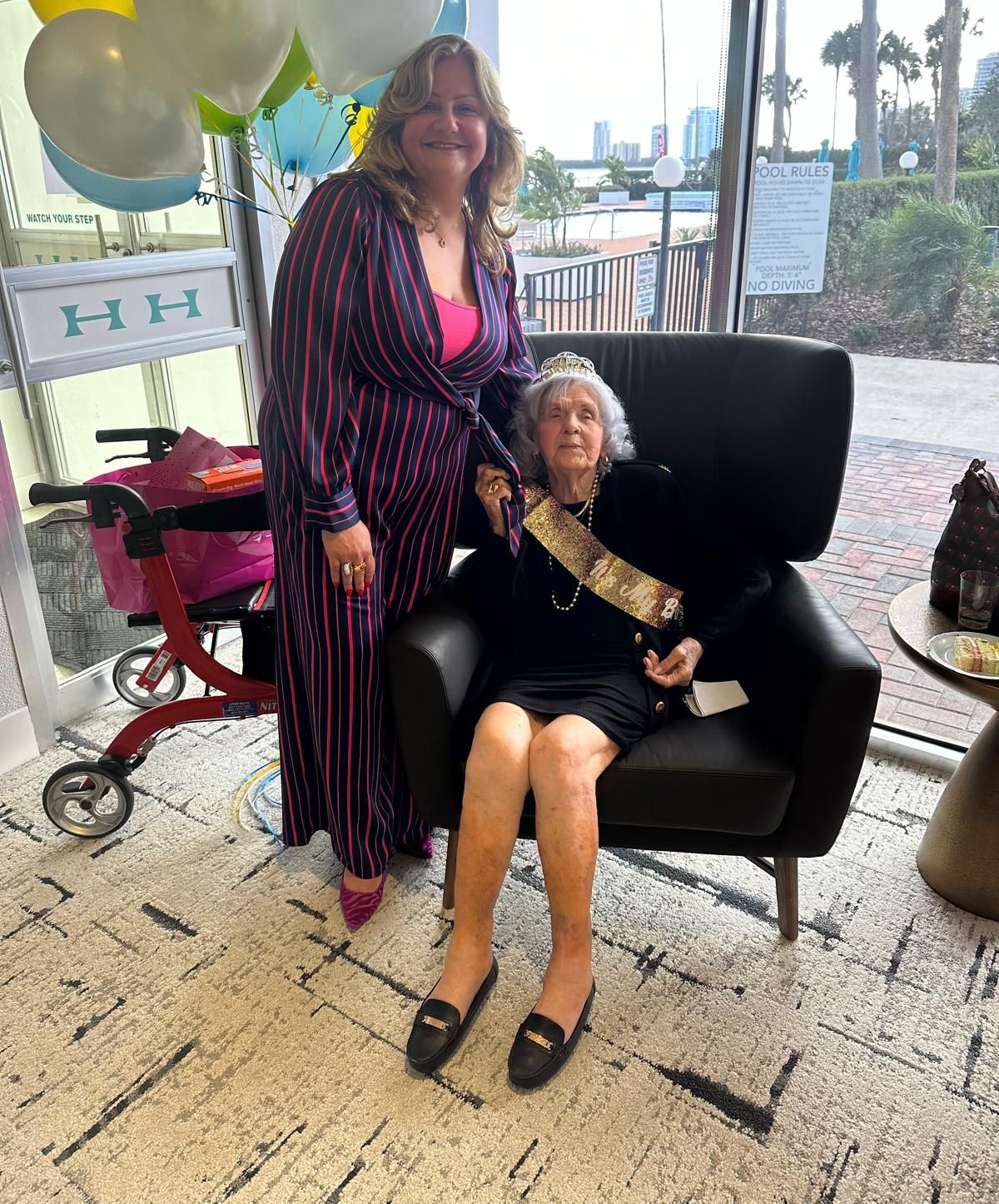 Woman in stripes stands with older woman in chair, holding a sash. Balloons, a walker, and a waterfront setting are visible.
