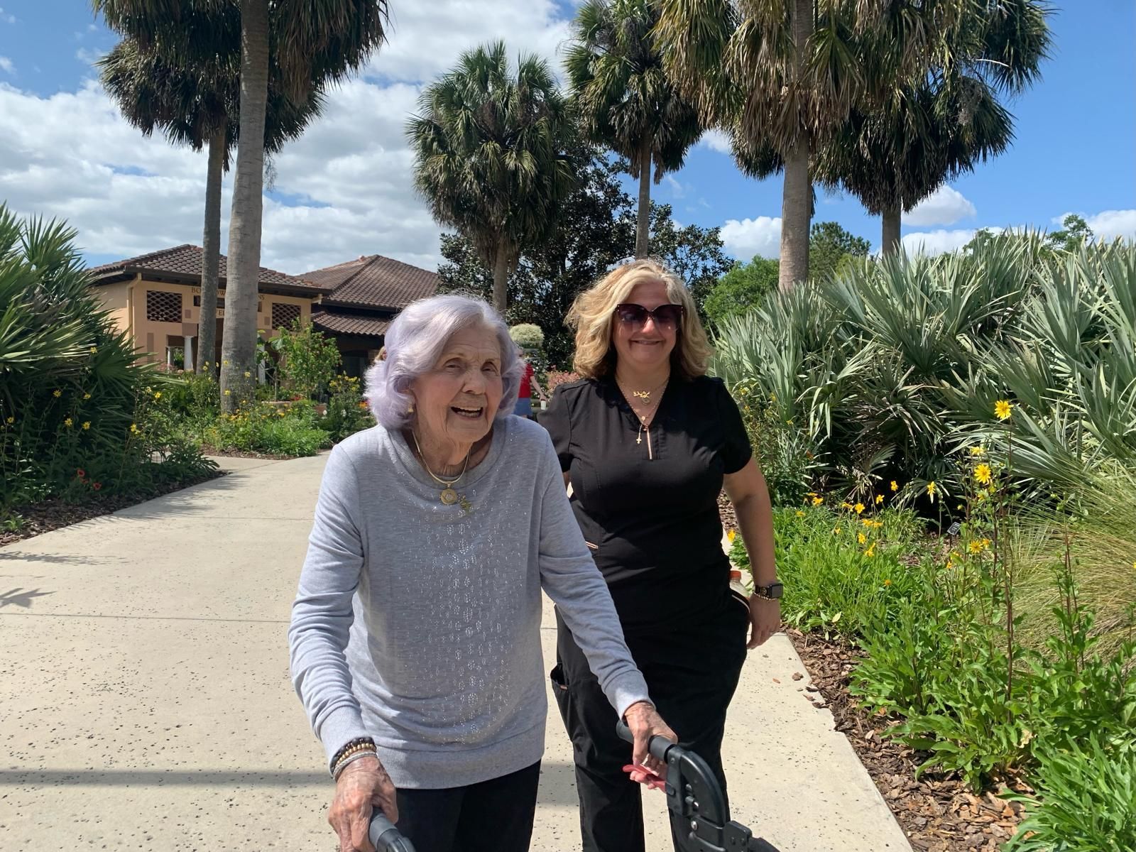 Woman using a walker walks outside with another woman on a path lined with palm trees and flowers.