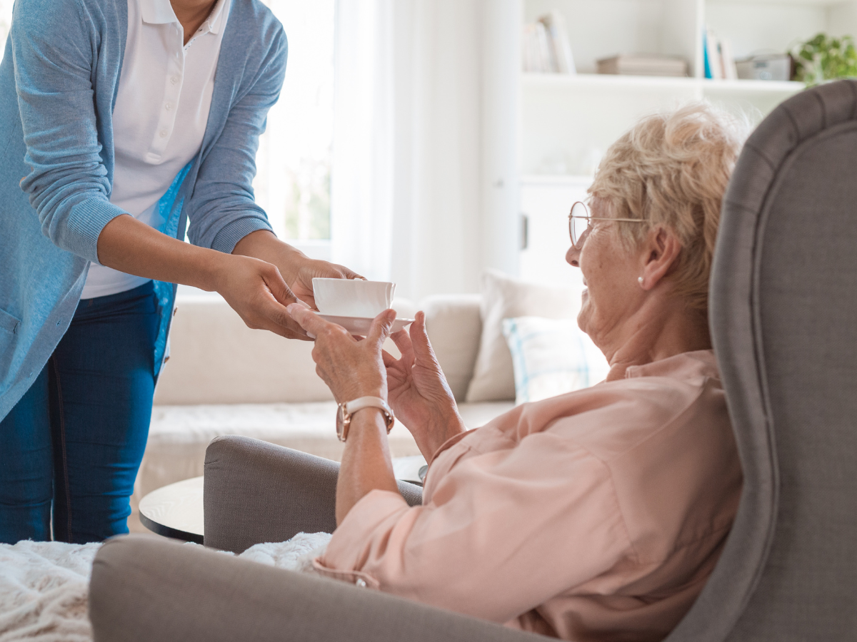 A person giving medication to another person seated in an armchair indoors.