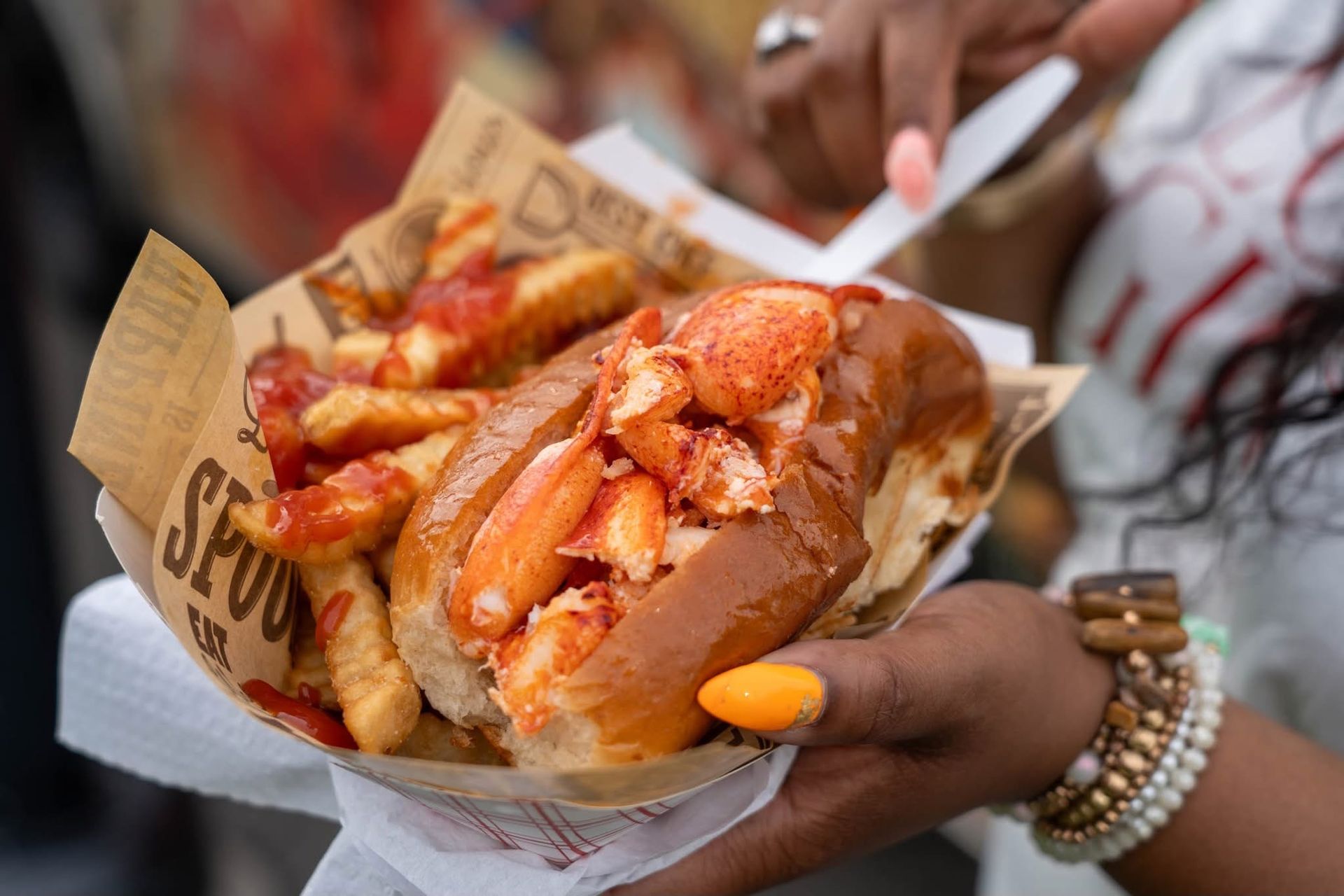 Hands holding a paper tray of fries and lobster rolls with sauce and lemon wedges