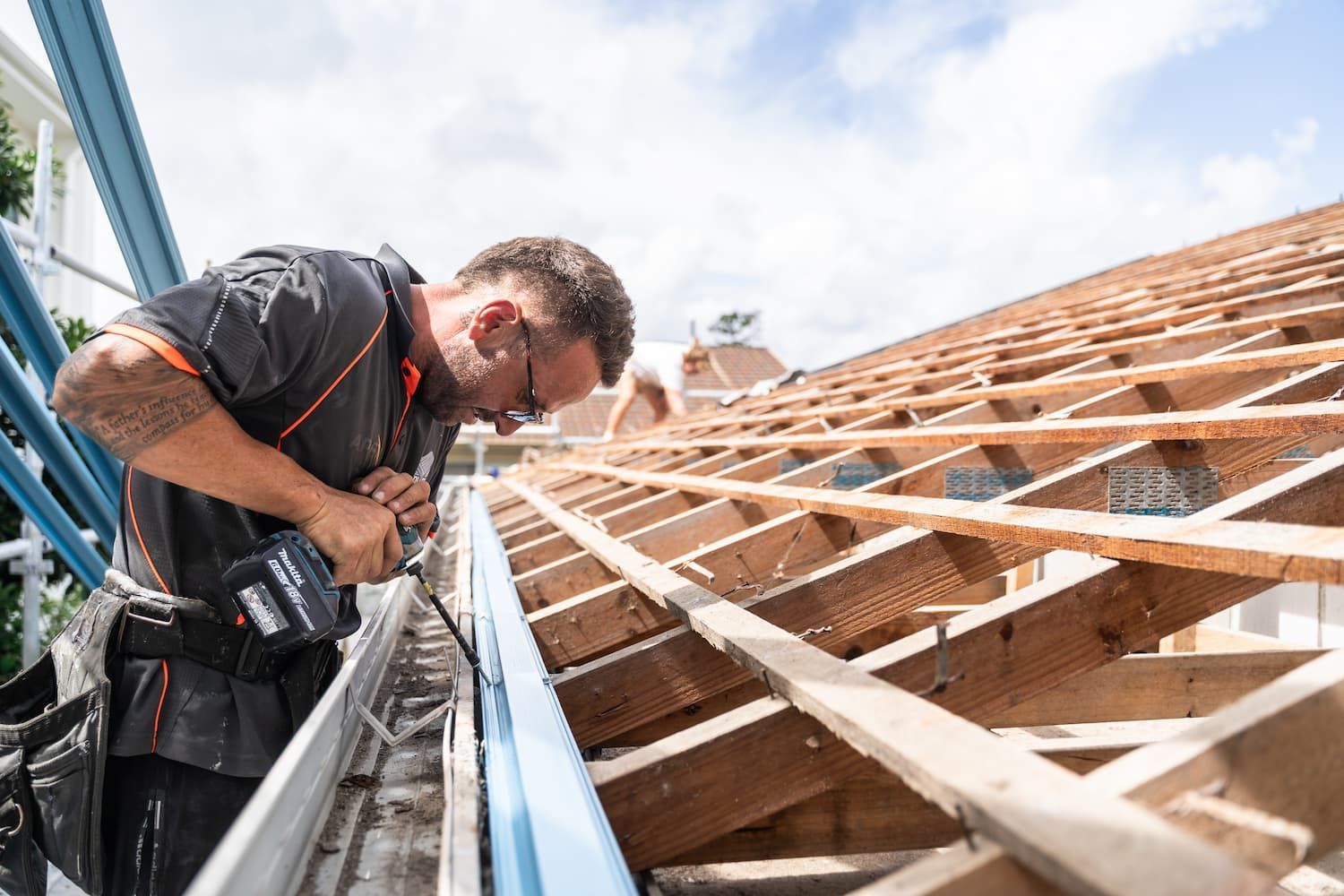 A worker wearing a tool belt uses a power drill to install a metal gutter on a wooden roof frame.