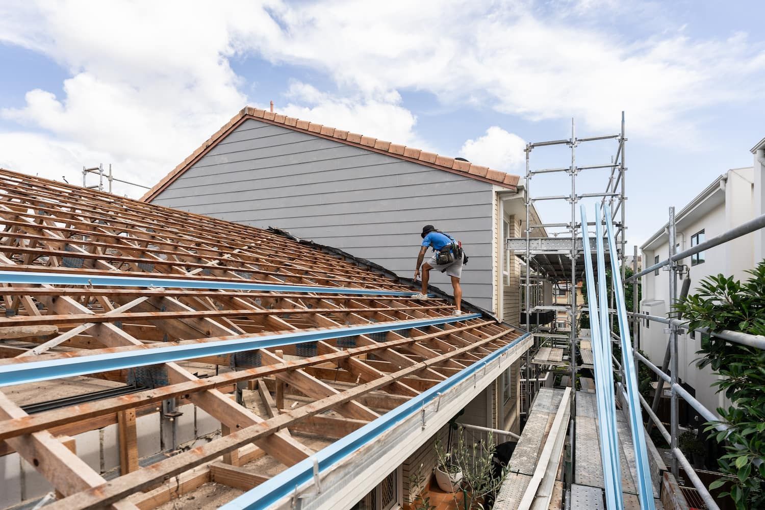 A worker stands on the wooden frame of a roof under construction, beside scaffolding on a bright, cloudy day.
