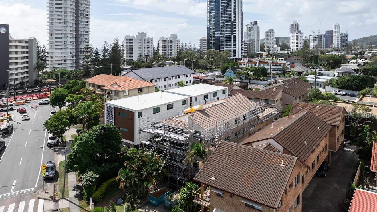 An aerial view shows low-rise residential buildings and a construction site surrounded by trees near a city skyline.