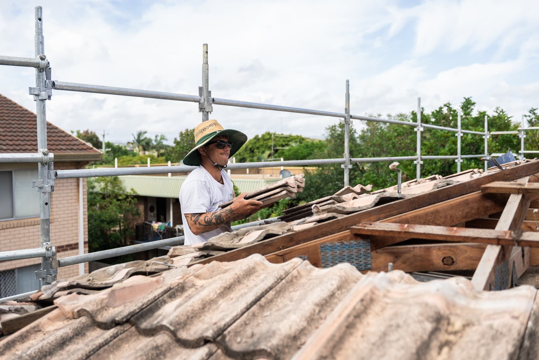 A person wearing a wide-brimmed sun hat works on a tiled roof framed by metal scaffolding during the day.