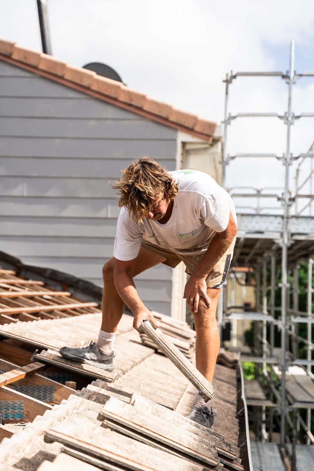 A person working on a roof, replacing or arranging light-colored roof tiles, with scaffolding visible nearby.