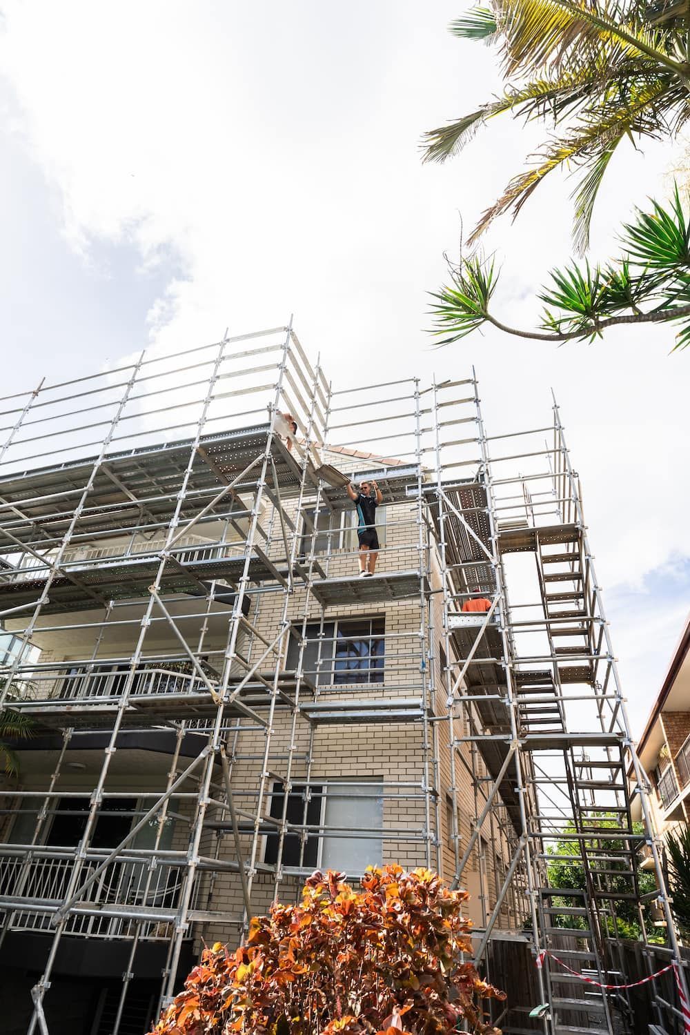 A person stands on metal scaffolding erected around a multi-story, beige-brick apartment building.
