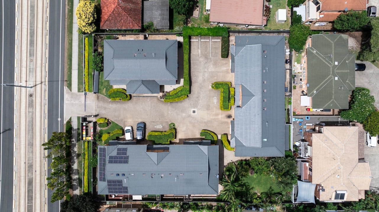 Aerial view of a complex with three grey-roofed buildings, a central paved courtyard, and surrounding greenery.