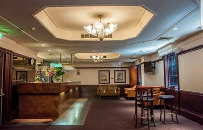 Interior of a bar with a patterned ceiling, seating, and a counter. Dark wood and warm lighting.