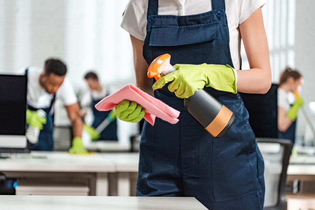 Cleaning staff in an office, wearing overalls and gloves, spraying and wiping desks, with other staff in the background.