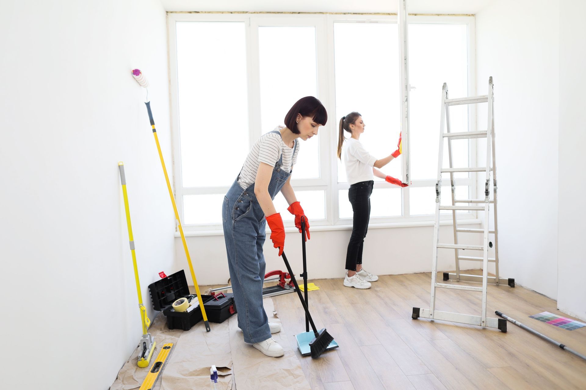 Two women cleaning a bright room with large windows. One sweeps; the other works near a ladder.
