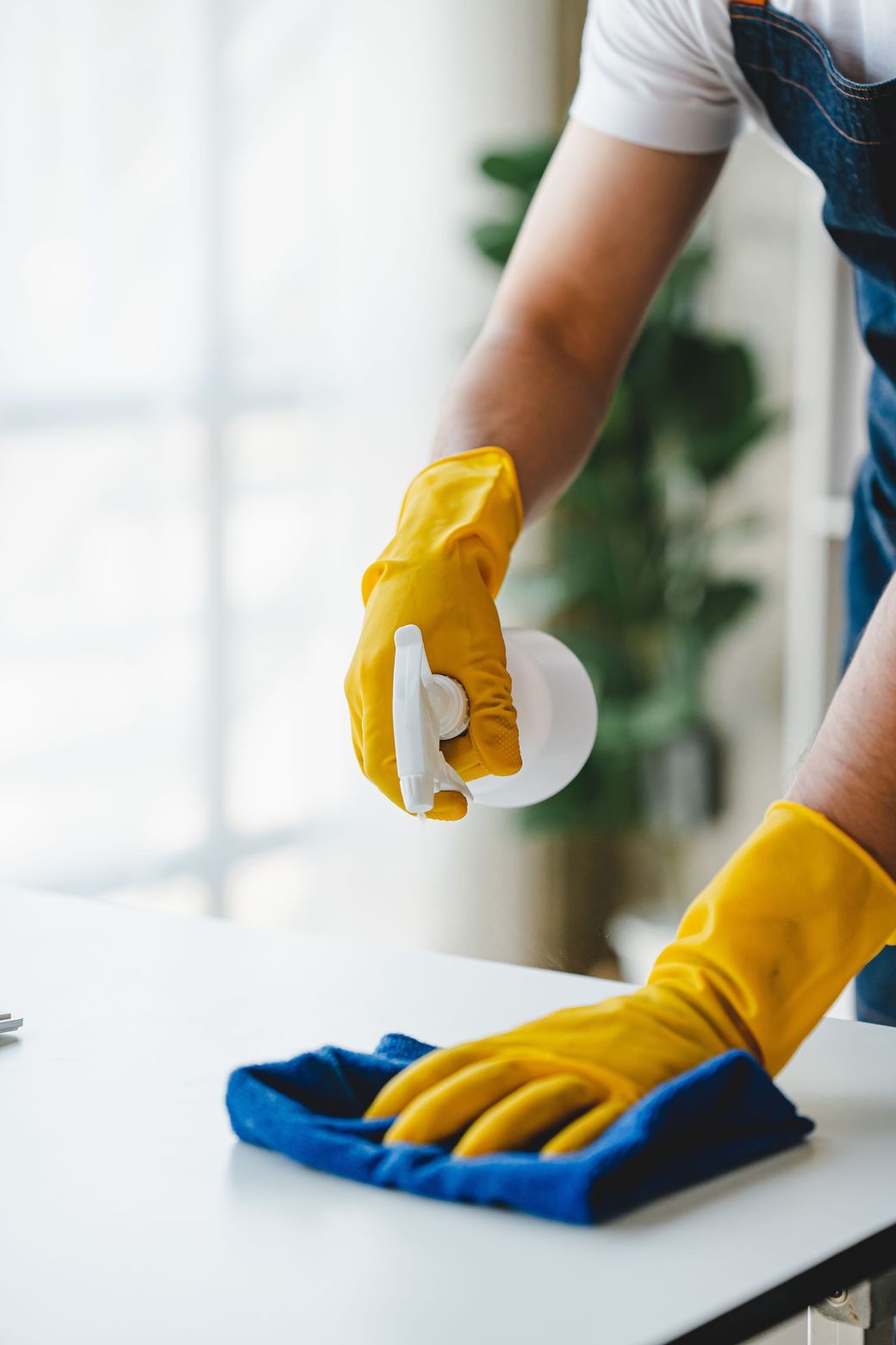 Person wearing yellow gloves sprays and wipes a white table with a blue cloth.