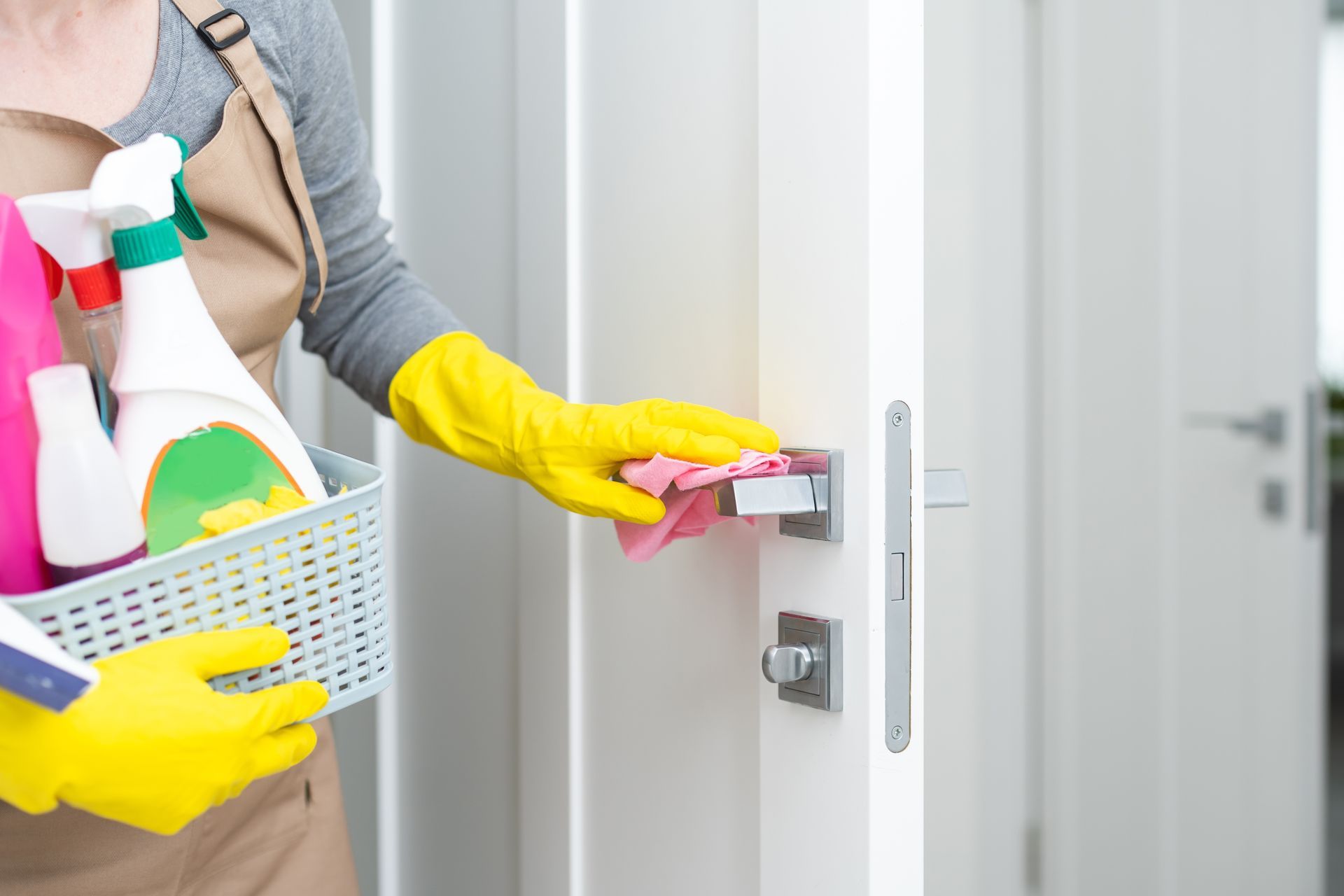 Person in yellow gloves cleaning a white door handle with a pink cloth, holding cleaning supplies.