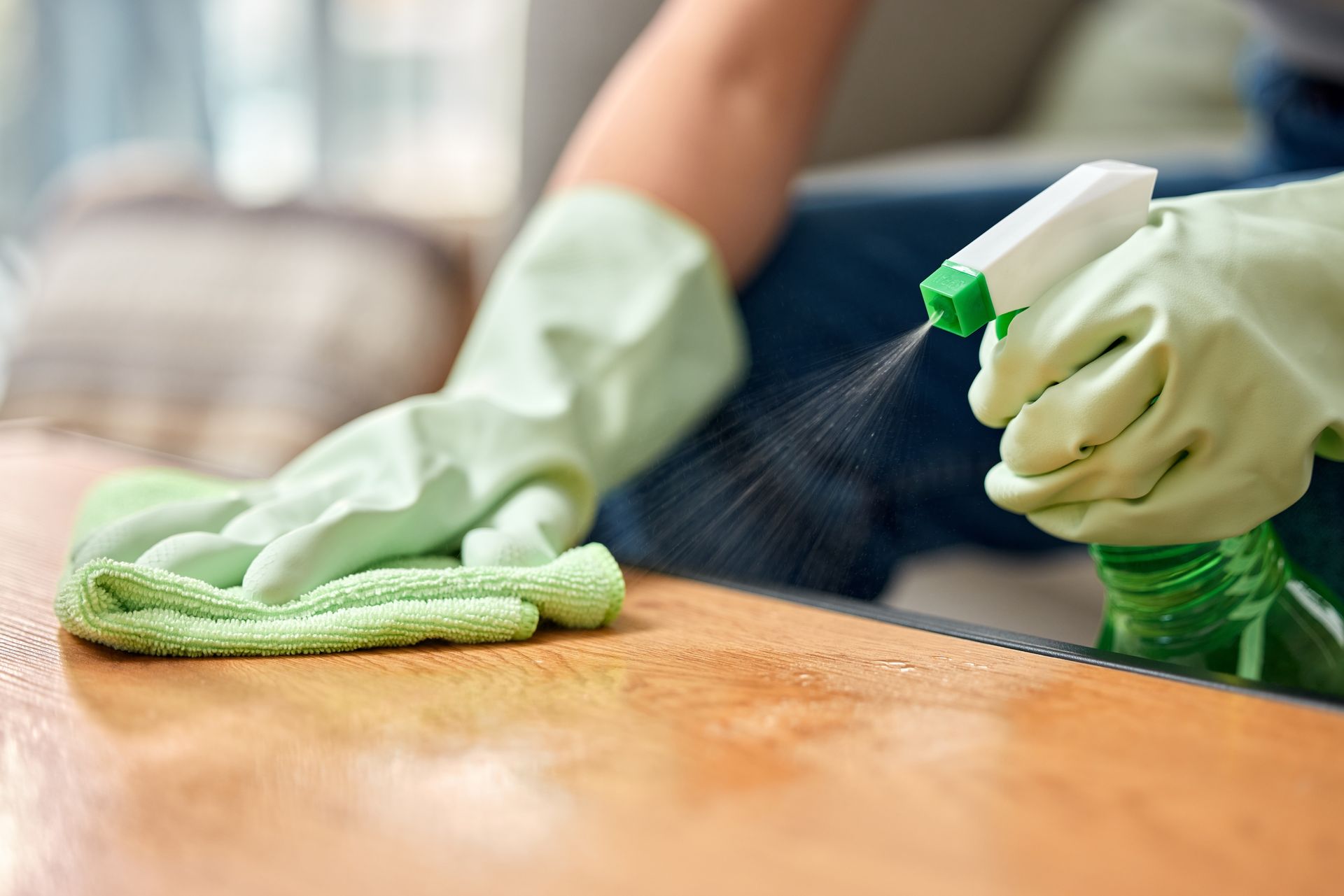 Person wearing green gloves spraying cleaner onto and wiping a wooden surface.