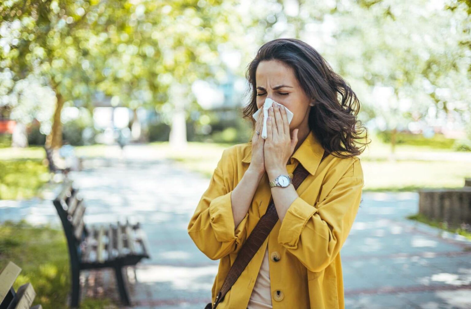 Woman in yellow jacket blowing her nose outdoors, representing whether allergies cause dry eyes