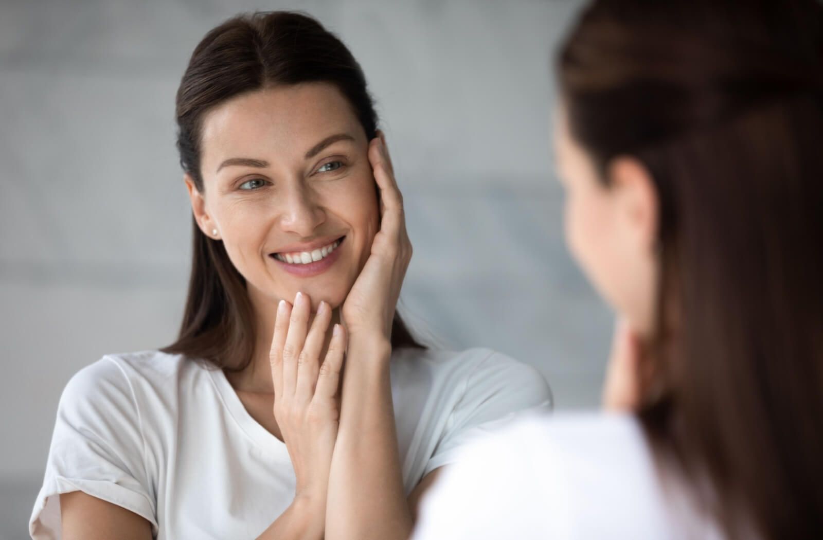 A woman in a white t-shirt looking in the mirror and touching her face. Represents whether IPL helps reduce wrinkles.