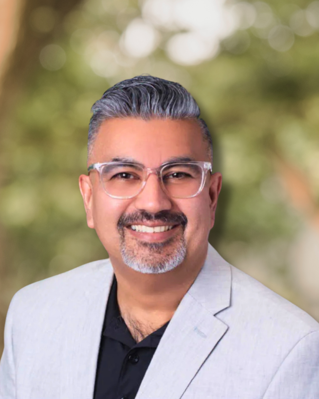 Man with graying hair, wearing glasses and a light blazer, smiling in front of a blurred green background.