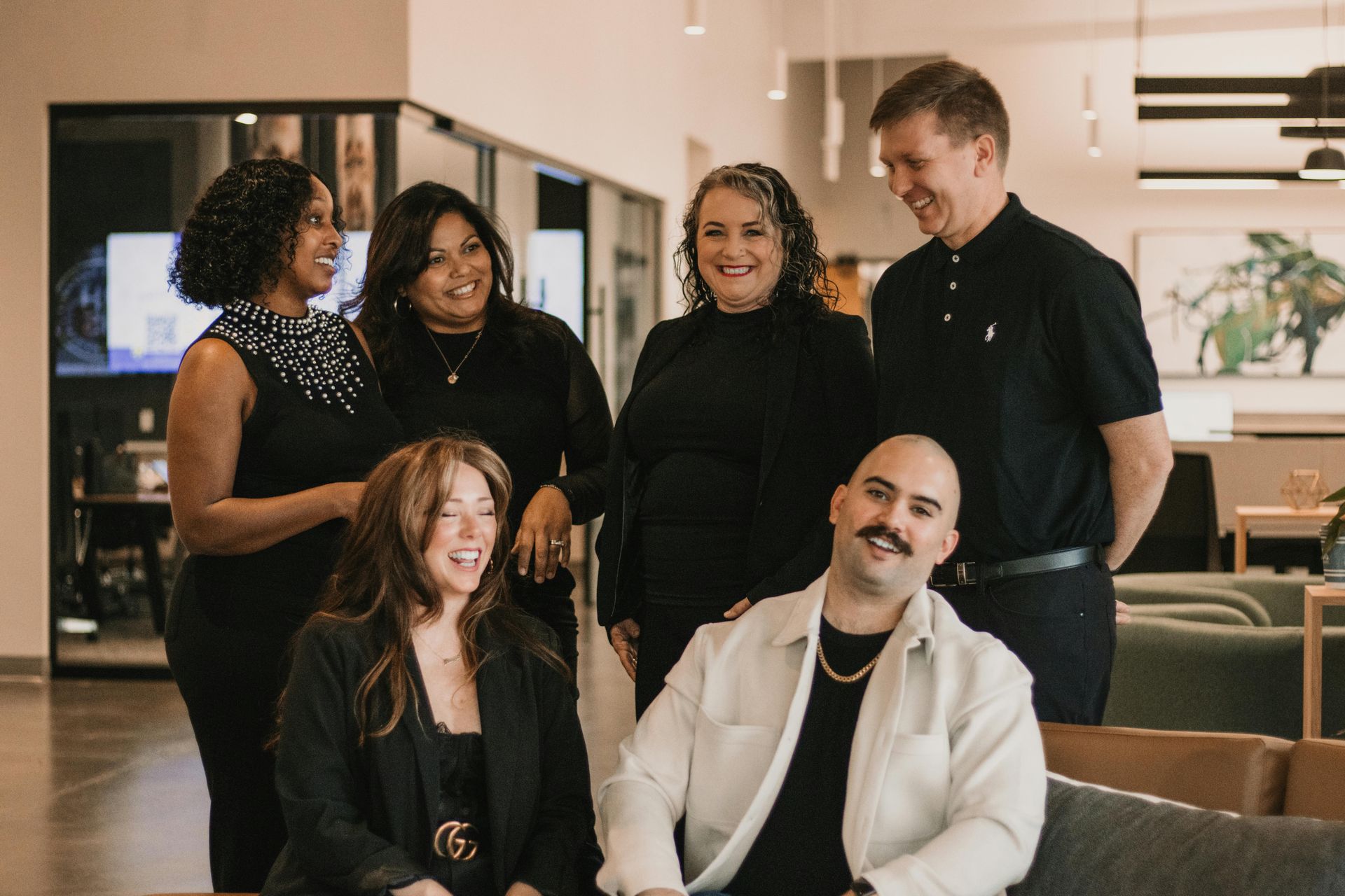Group of six people smiling in an office setting, some standing, some seated.