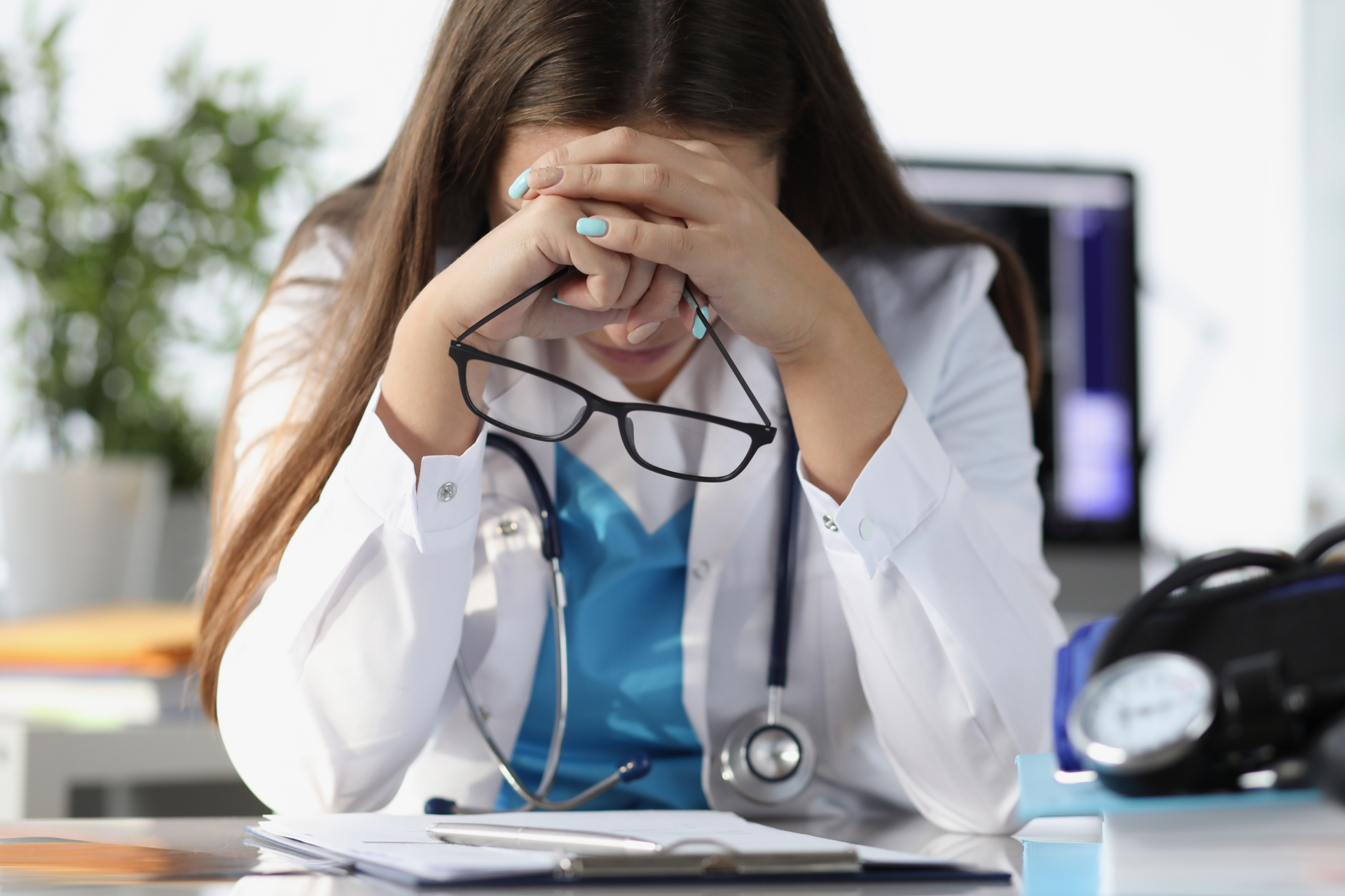 A medical professional holding her glasses while pressing her head to her arms in apparent frustration.