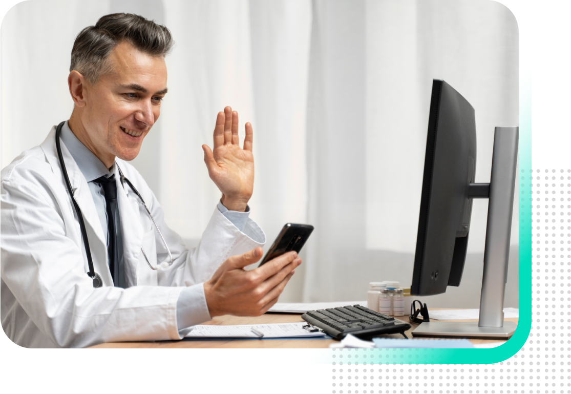 A doctor is sitting at a desk using a computer and a cell phone.