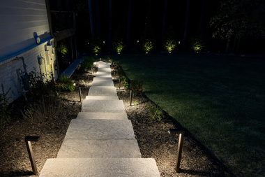 Lit walkway beside a building at night, lined with small path lights and leading into a dark lawn