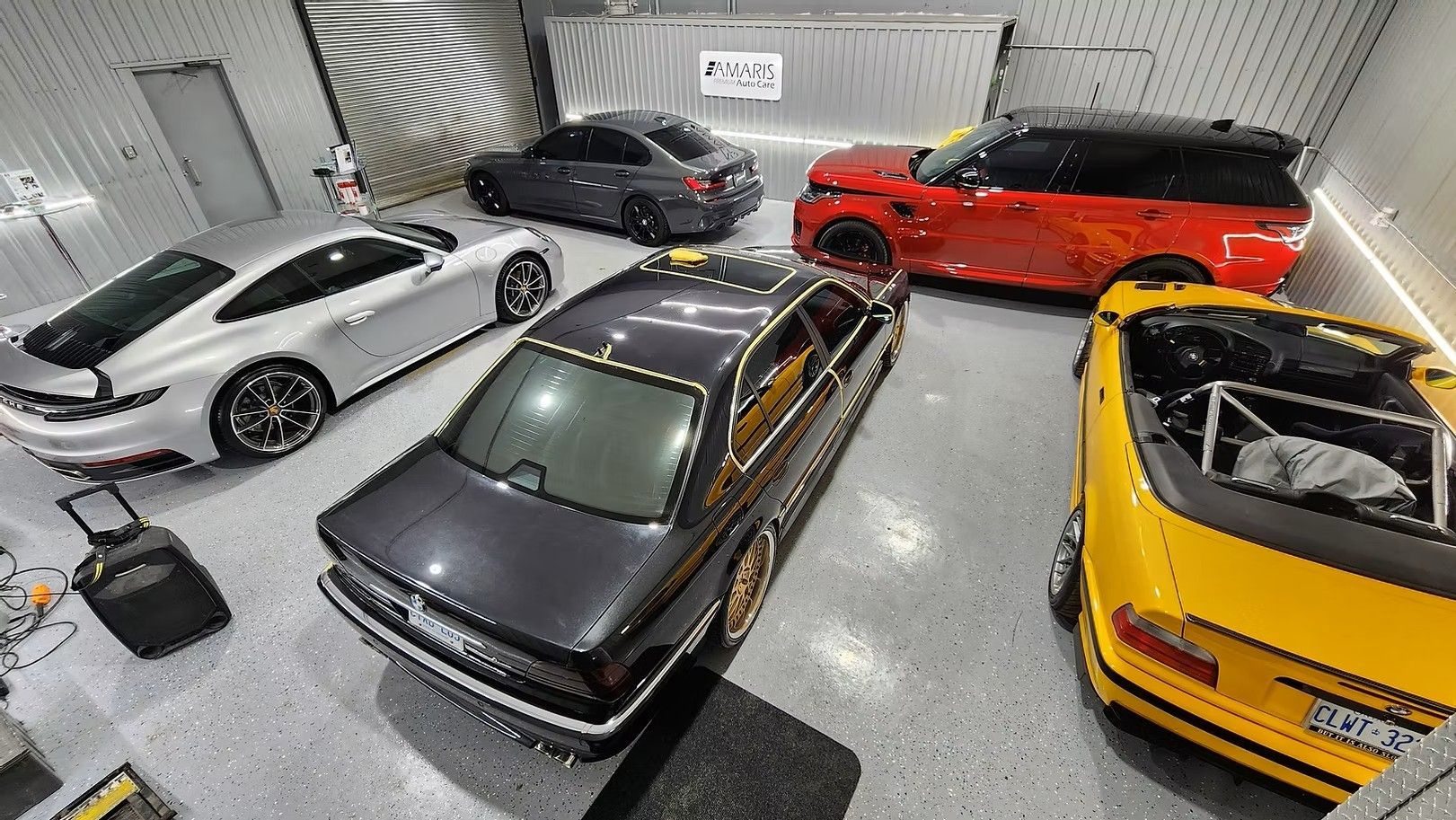 Six luxury cars in a garage, including white Aston Martin, red Range Rover, and yellow convertible.