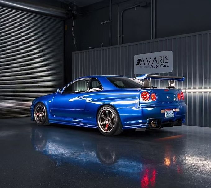 Blue Nissan Skyline R34 sports car with a large spoiler in a garage, reflecting on the glossy floor.