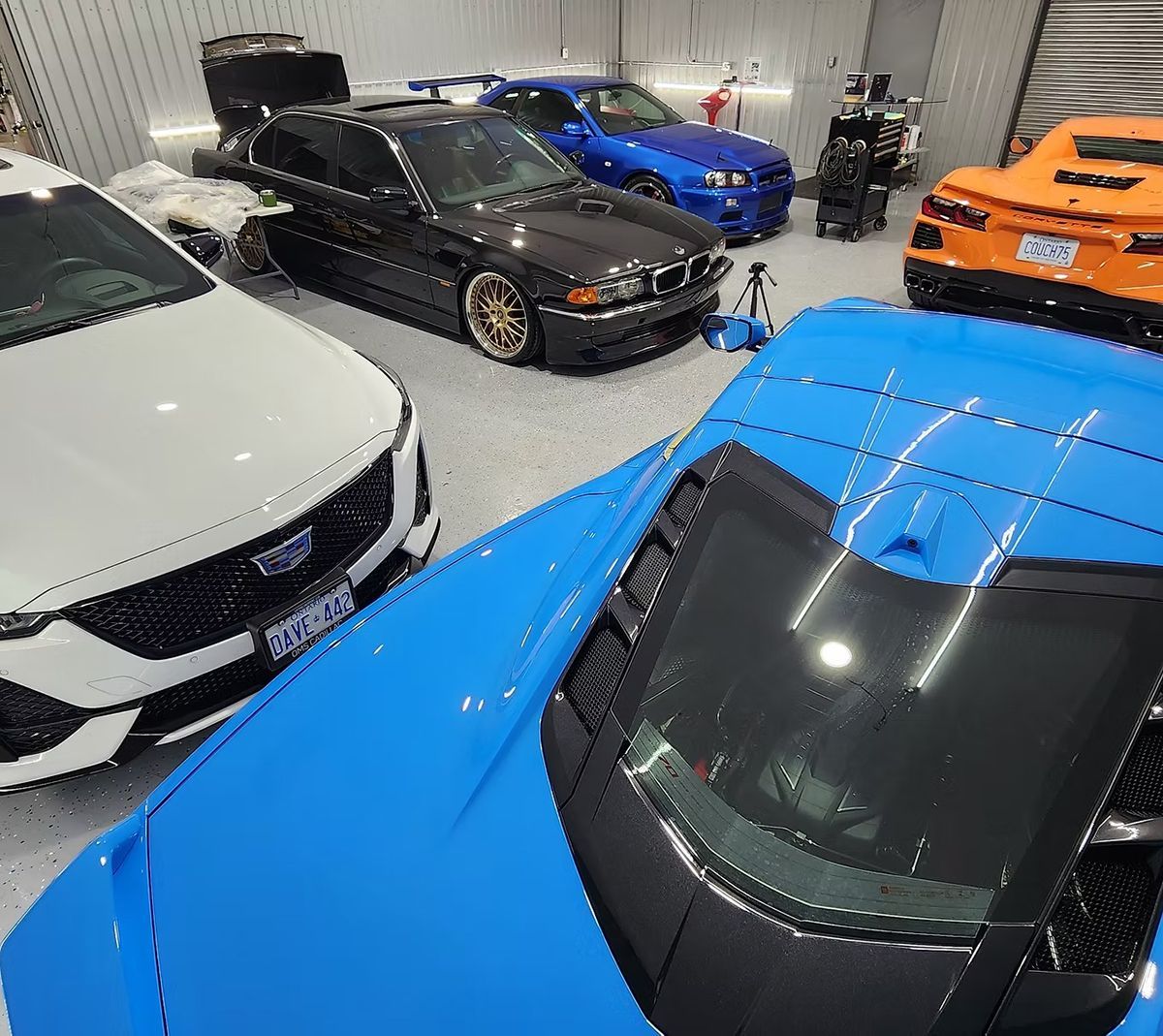 Cars in a garage: blue, white, black, and orange vehicles parked. Shop interior, bright lighting.
