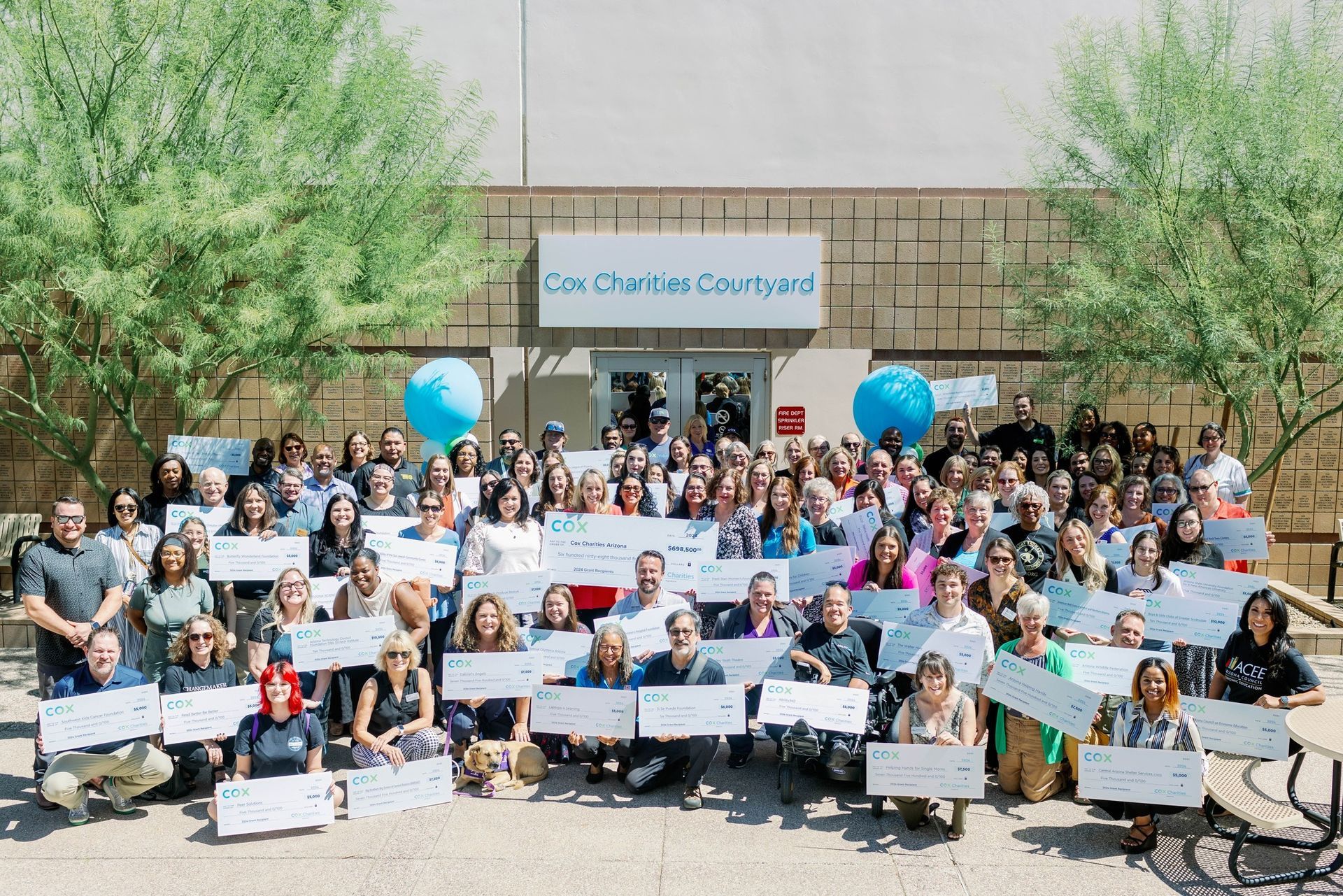 Large group of people holding checks outside a building. Blue balloons, sunny day.