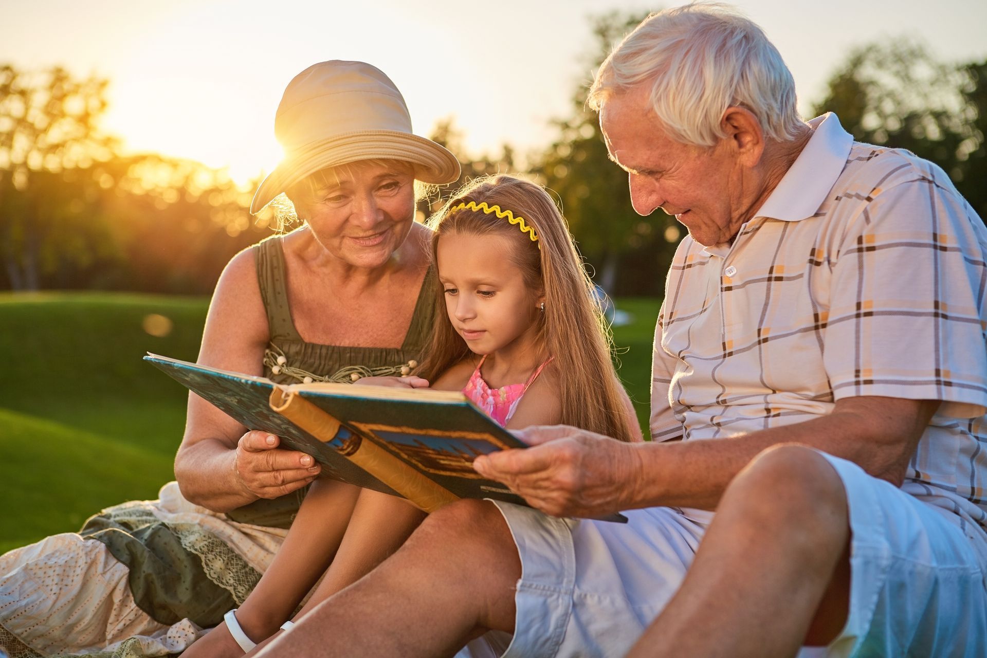 Grandparents and granddaughter reading a book together on a grassy hill in the sunlight.