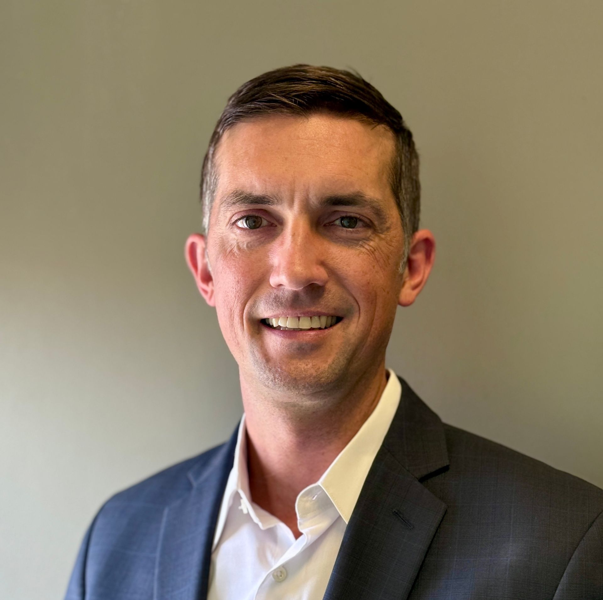 Man in a dark suit jacket, white shirt, smiling, looking at the camera against a gray background.